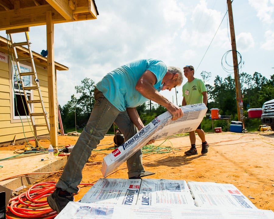 A older man carries shingles as volunteers prepare to replace a roof.