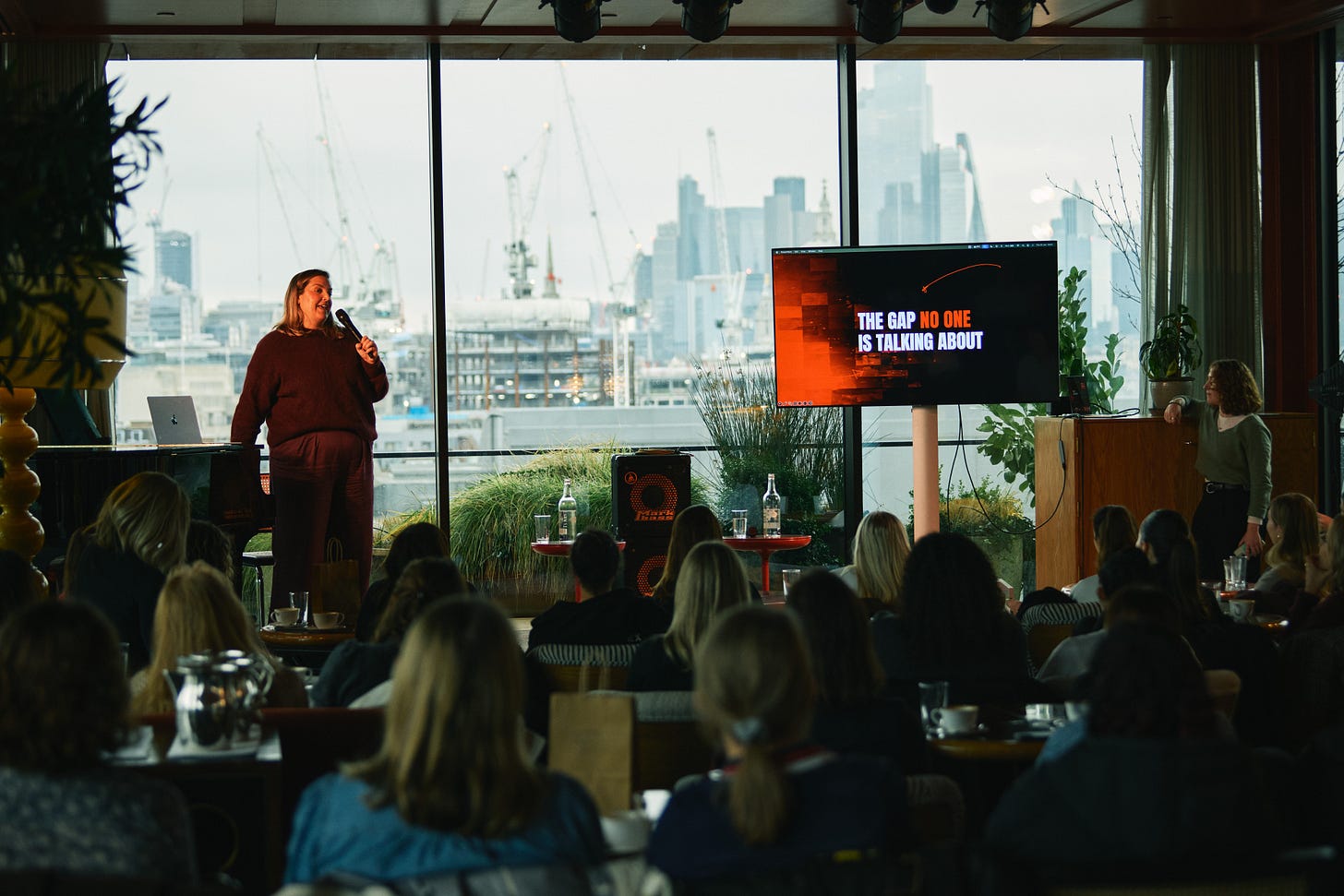 Photo of Alice from Charlton FC, on stage at Soho House with a microphone in her hand. Behind her is a window showing the London skyline. Next to her is a screen with red Charlton branding on it that says "the gap no one's talking about". In the foreground of the photo are the backs of the audience's heads.