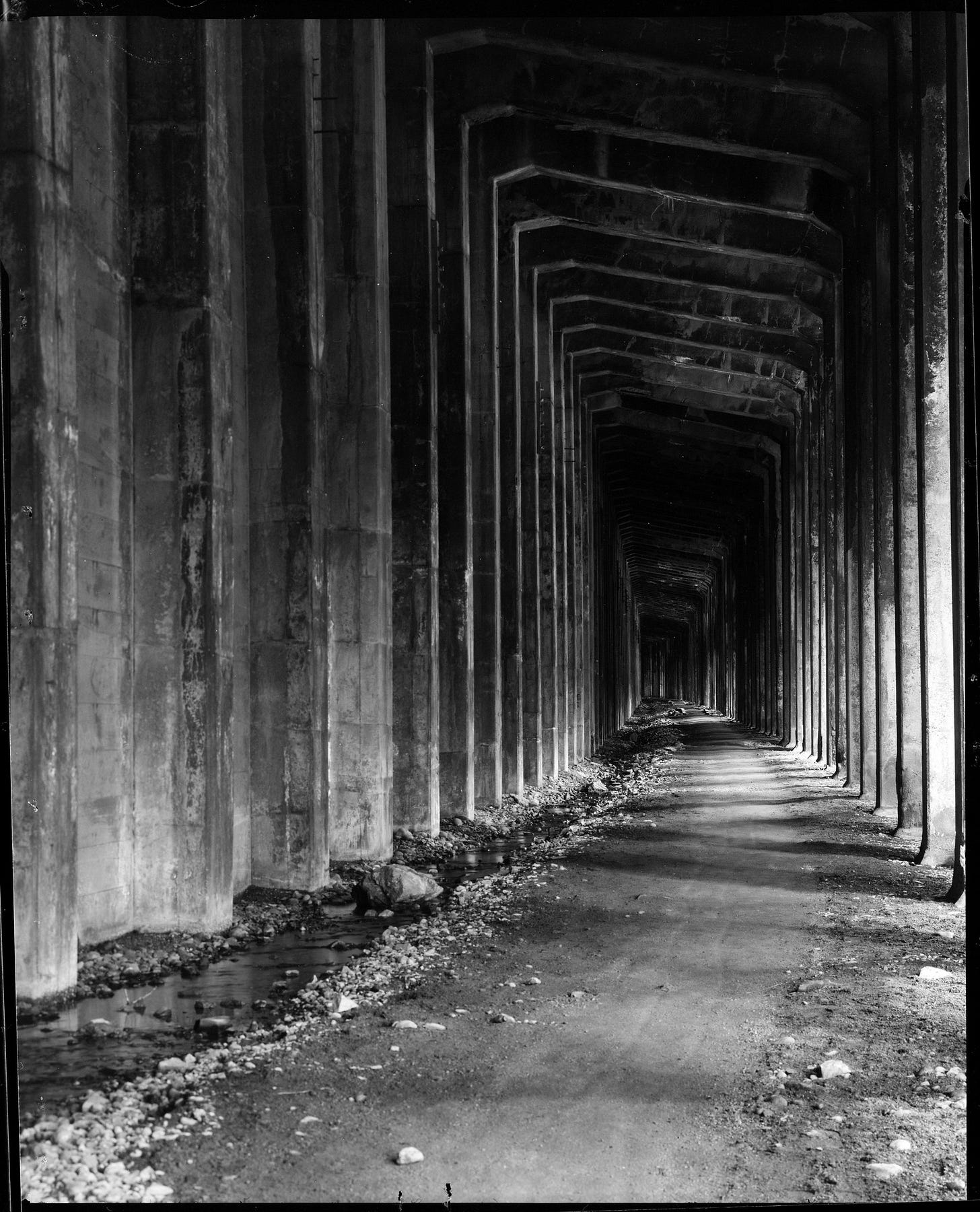 Tall columns and pillars line what used to be a railroad grade. The rails have been removed, leaving a packed earth path moving through a concrete snowshed. 