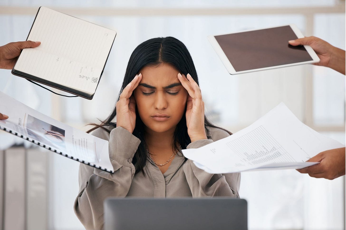 Stressed out and burnt out business lady. She is sat at her computer holding her head while lots of hands hold business related things at her.