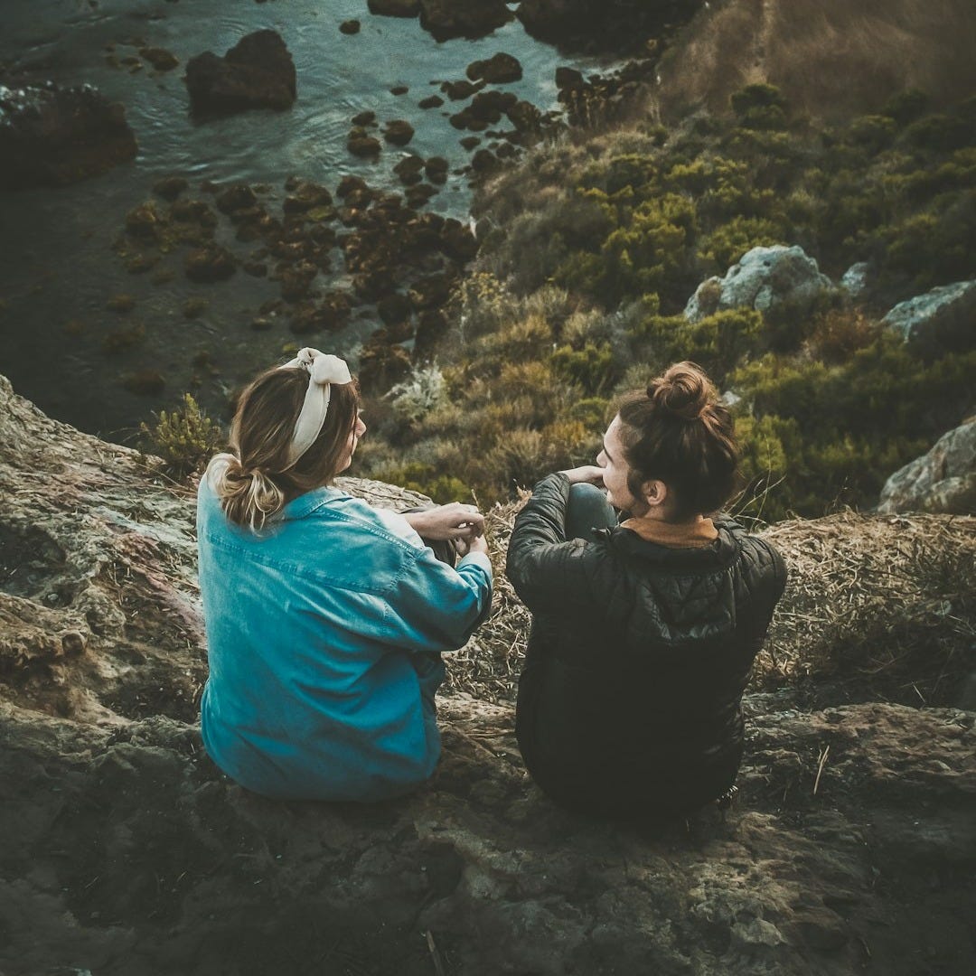 two women sitting on cliff while chatting viewing blue body of water
