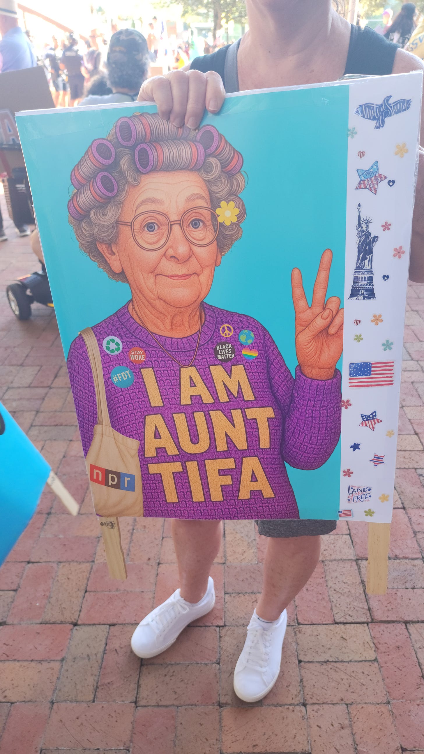 A person holds a colorful sign showing an illustration of an elderly woman flashing a peace sign with the text "I AM AUNT TIFA."