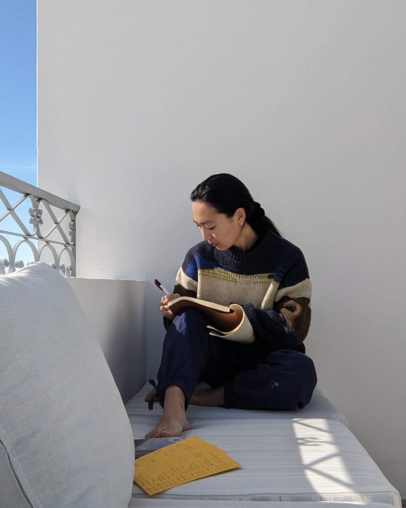 Photo of Asian person sitting outside on a sofa and writing in their notebook, in the sunshine.