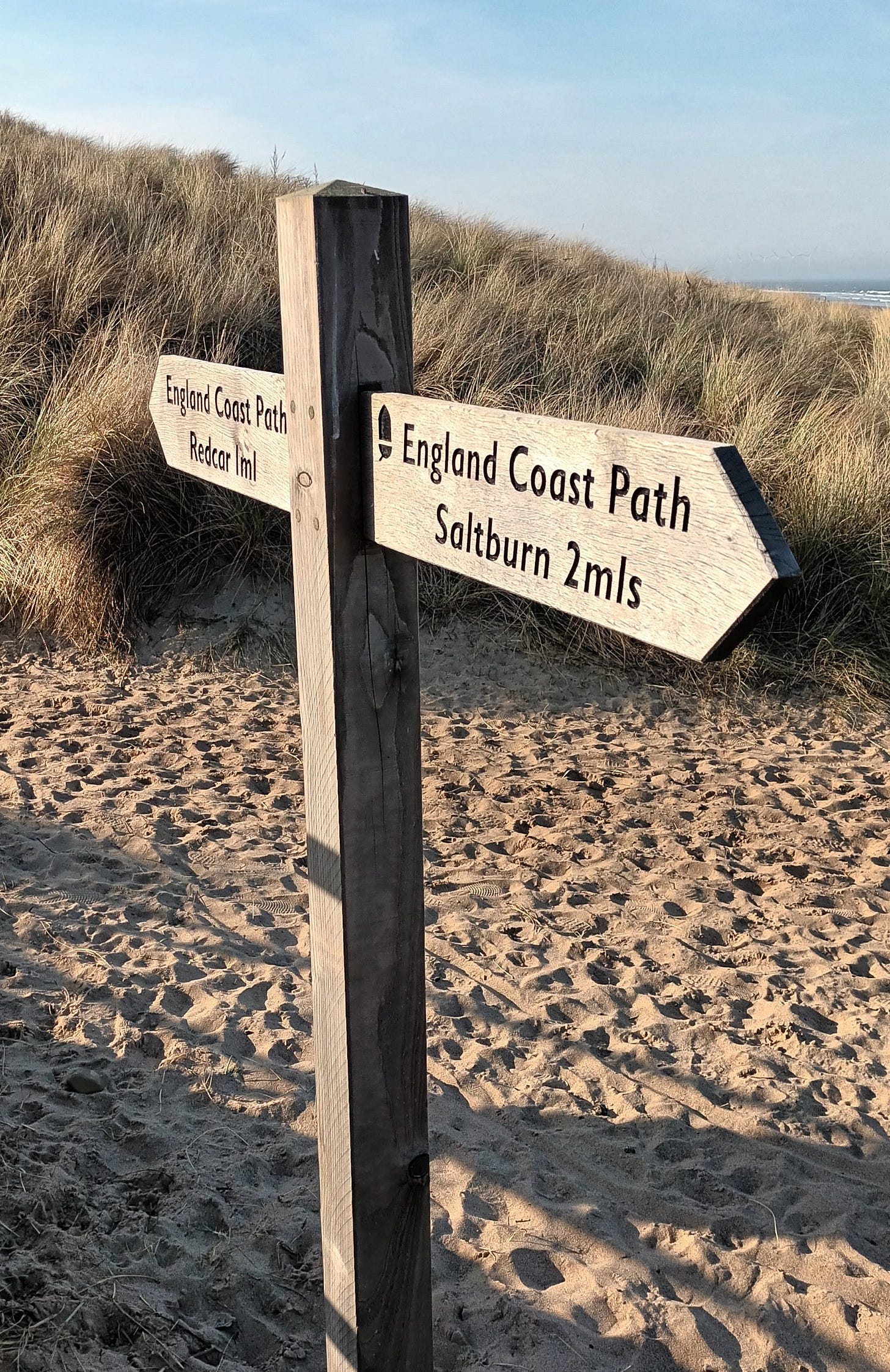 England Coast Path sign, Redcar 1m, Saltburn 2m, sand below, dunes behind and blue sky above