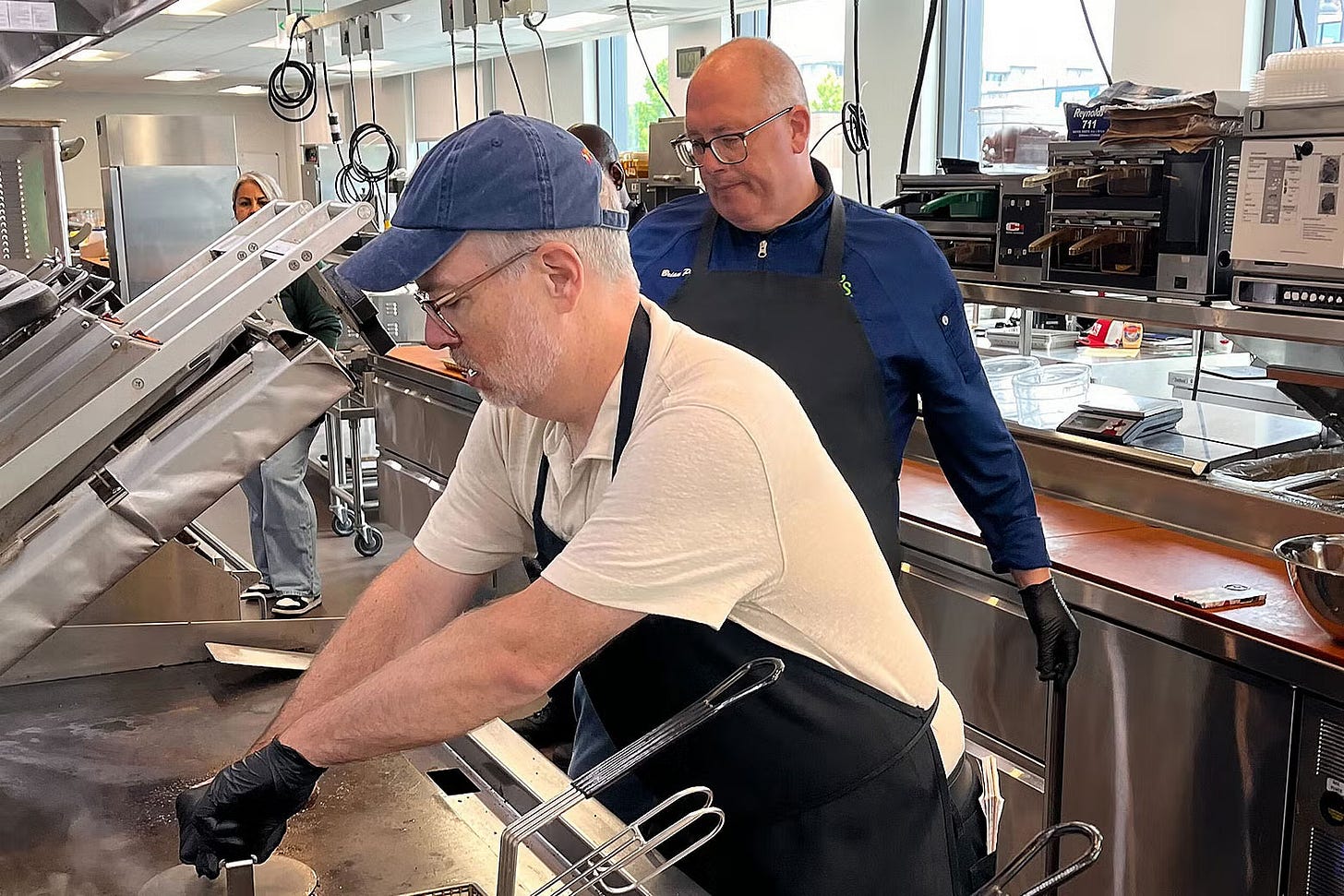 Two men inside a kitchen, with one cooking a burger. Two men inside a kitchen, with one cooking a burger.