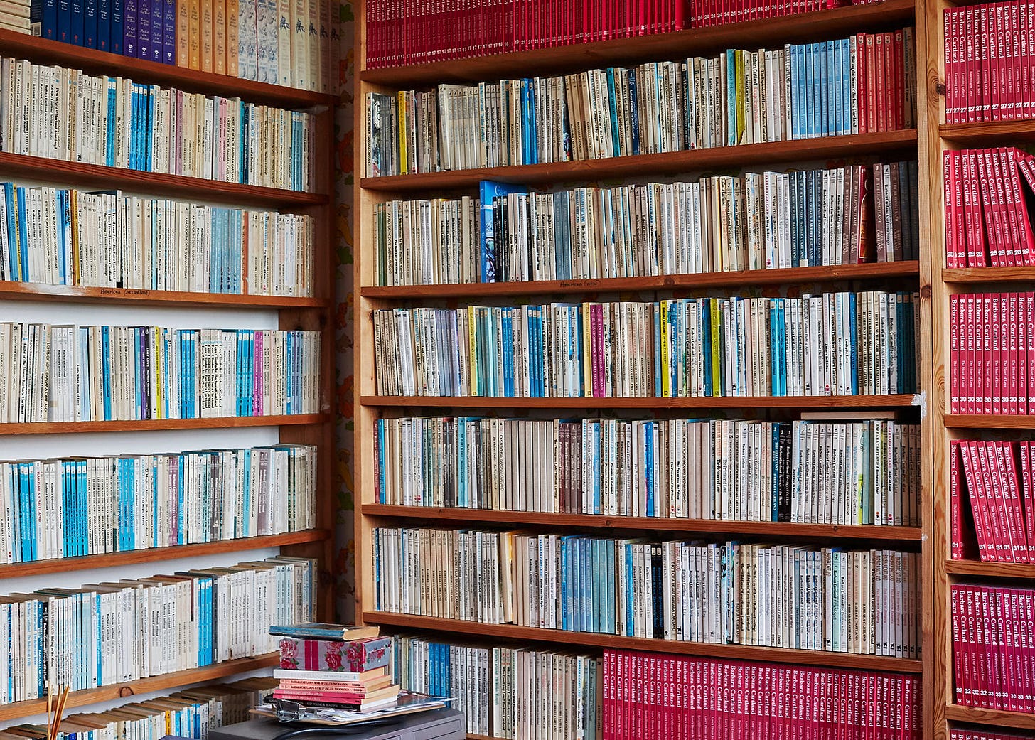 Bookshelves filled with books of varied colors, mostly reds and blues, in a cozy library. A small stack of books sits on a nearby table. Barbra Cartland Claudia Legge 