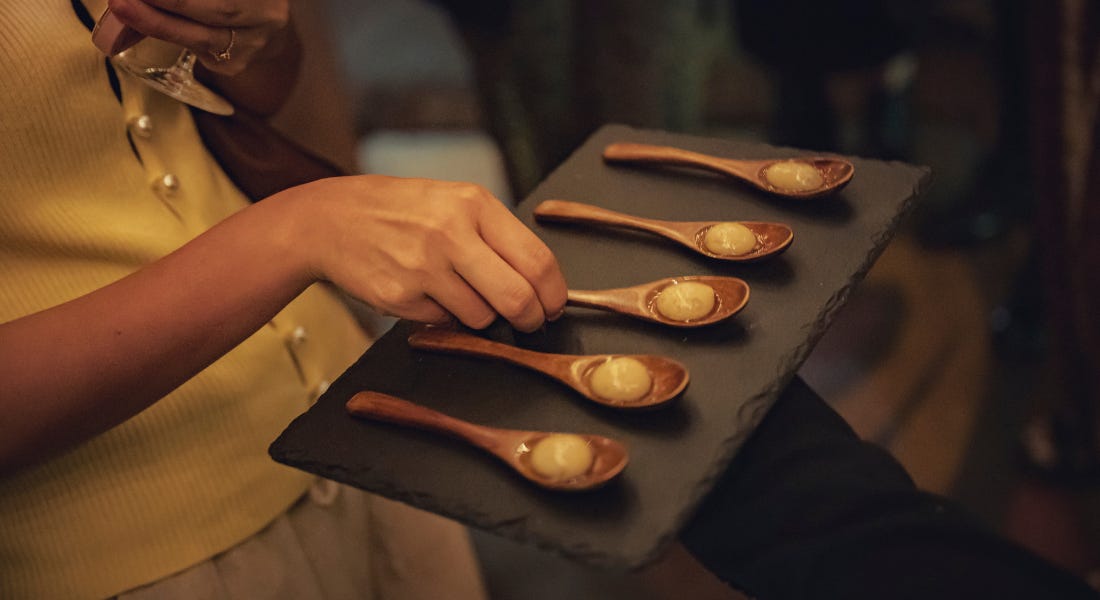 A close-up of five wooden spoons with olive shaped entrees on top of them on top of a black slate, with a person picking them up to the left A close-up of five wooden spoons with olive shaped entrees on top of them on top of a black slate, with a person picking them up to the left