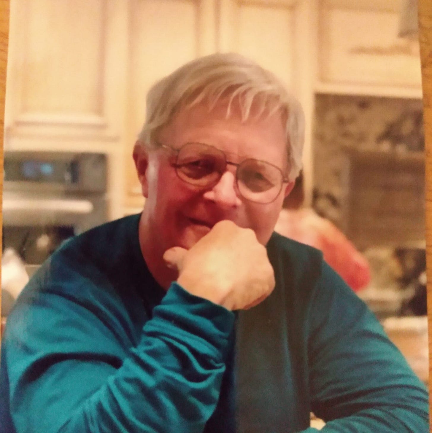 Bruce Frendahl seated indoors, wearing glasses and a long-sleeve teal shirt, resting his chin on his hand and smiling gently. A softly lit kitchen is visible in the background.