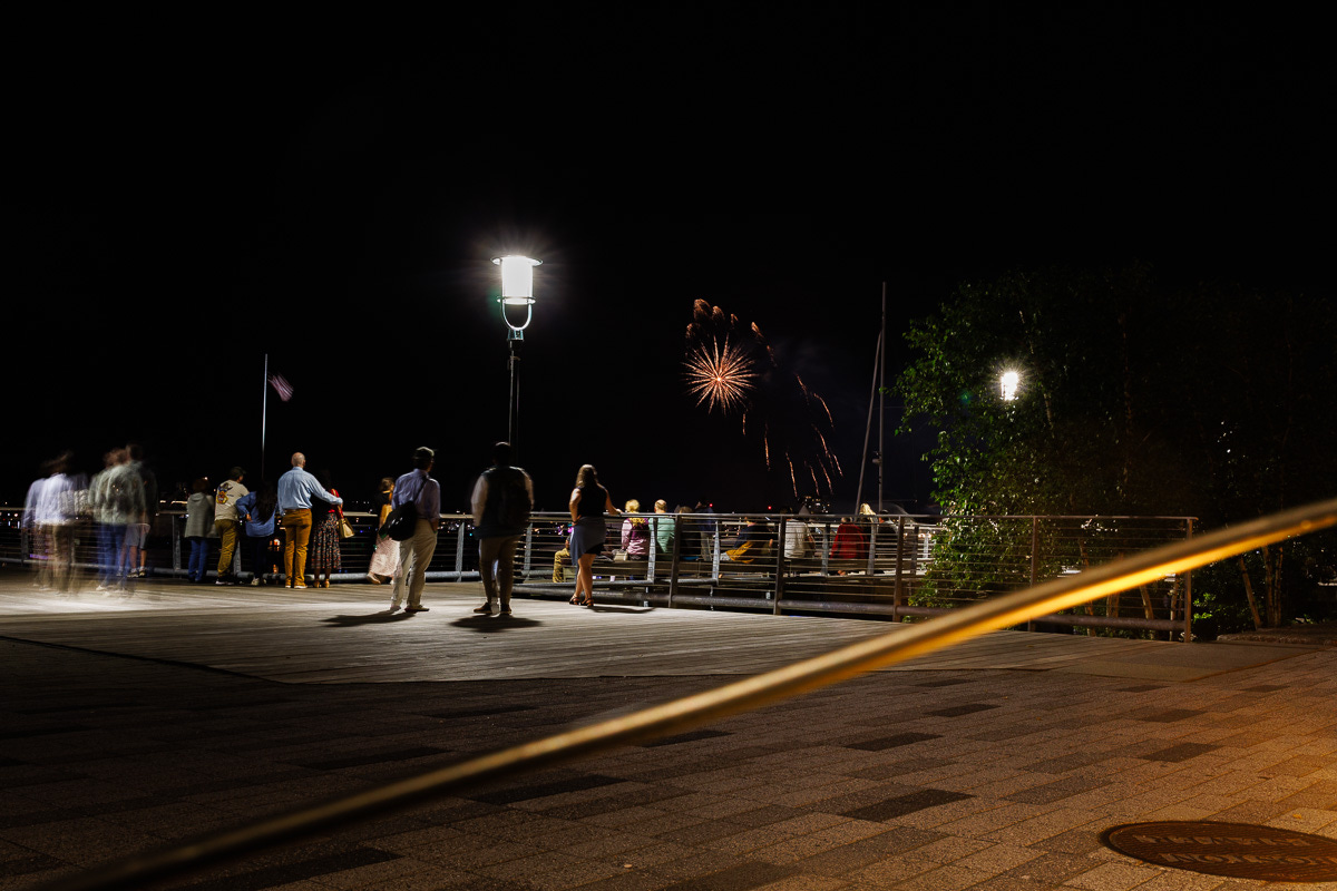 Fireworks bursting over Boston Harbor with blurred silhouettes of people watching along the pier