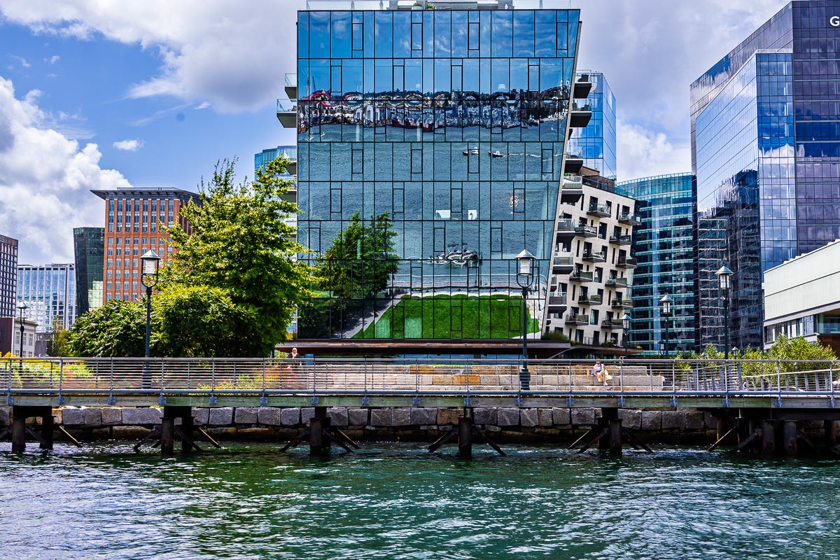 Modern glass building in Boston Seaport reflecting water, boats, and photographer’s boat in the facade