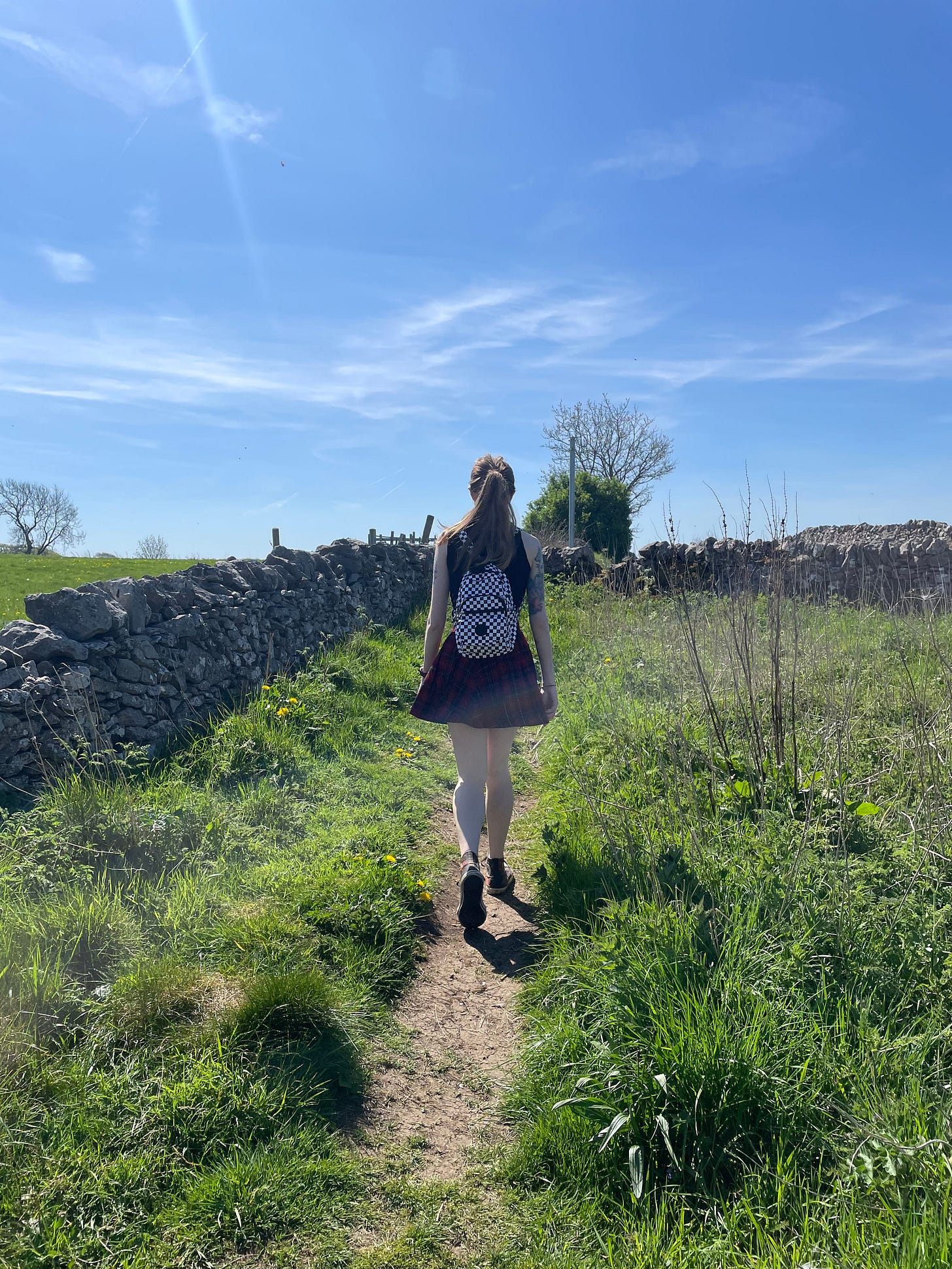 The writer, with their back to the camera, walking up a grassy hill. They are wearing a black, sleeveless t-shirt, tartan skirt and Converse. They have a black and white checked backpack on their back. The top of the photo is the sky which is incredibly blue. 