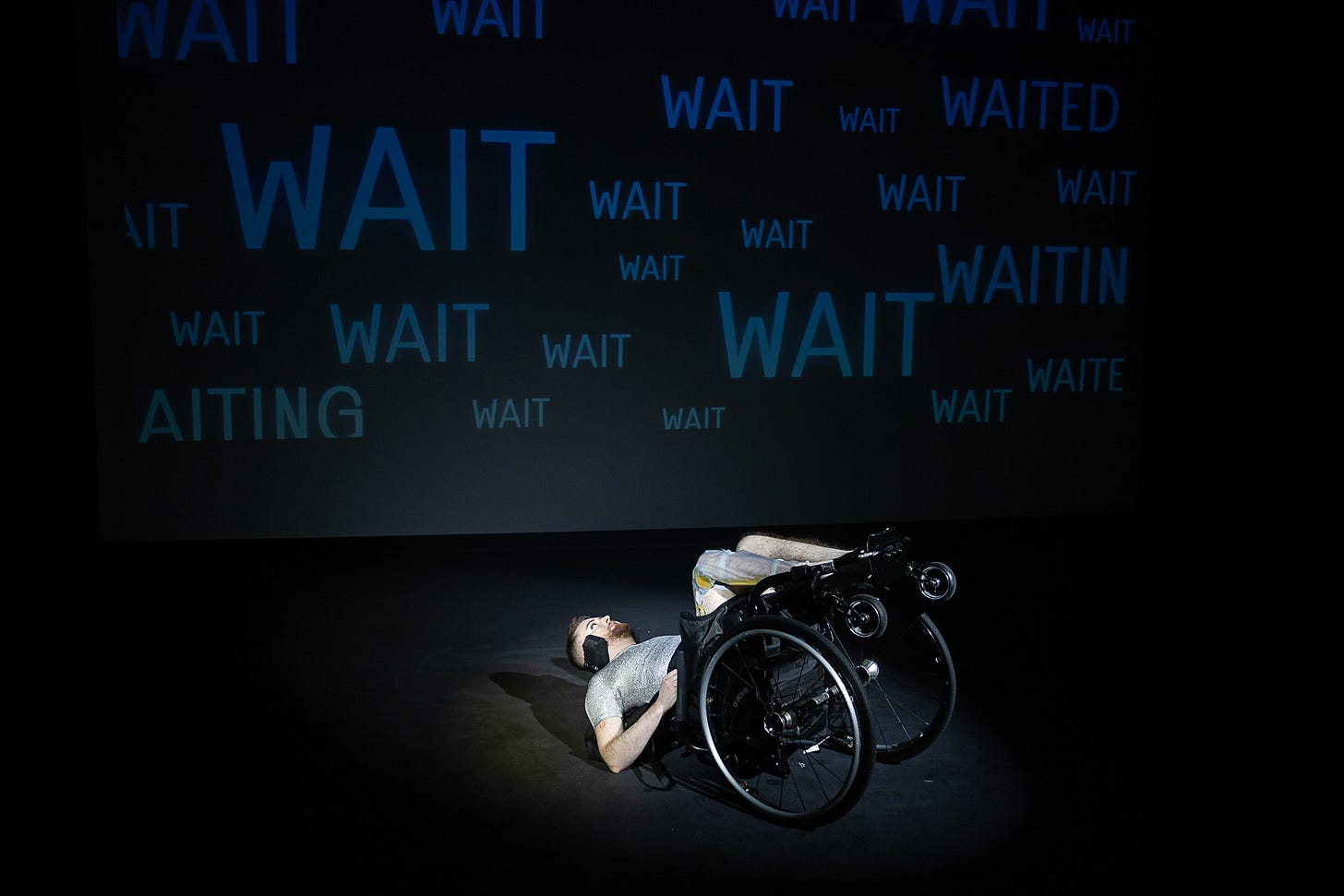 Image: Jamie Hale, a white person with dark red hair and a ginger beard lies in a manual wheelchair tipped onto its back on the stage. Behind them is the word WAIT projected in navy onto a black background over and over again.