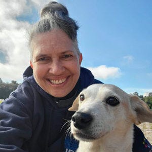 A imagem mostra uma mulher sorrindo, em um ambiente ao ar livre, durante o dia. Ela parece estar tirando uma selfie. A mulher tem cabelo grisalho preso no alto da cabeça e usa um moletom escuro. Ao lado dela, bem próximo do rosto, está um cachorro de porte pequeno a médio, com pelagem clara, predominantemente branca, e orelhas caídas. O cachorro olha levemente para o lado, enquanto a mulher olha diretamente para a câmera, transmitindo uma expressão de alegria e carinho. Ao fundo, há um céu azul com algumas nuvens e uma paisagem natural desfocada, que sugere um campo ou área aberta. A imagem mostra uma mulher sorrindo, em um ambiente ao ar livre, durante o dia. Ela parece estar tirando uma selfie. A mulher tem cabelo grisalho preso no alto da cabeça e usa um moletom escuro. Ao lado dela, bem próximo do rosto, está um cachorro de porte pequeno a médio, com pelagem clara, predominantemente branca, e orelhas caídas. O cachorro olha levemente para o lado, enquanto a mulher olha diretamente para a câmera, transmitindo uma expressão de alegria e carinho. Ao fundo, há um céu azul com algumas nuvens e uma paisagem natural desfocada, que sugere um campo ou área aberta.