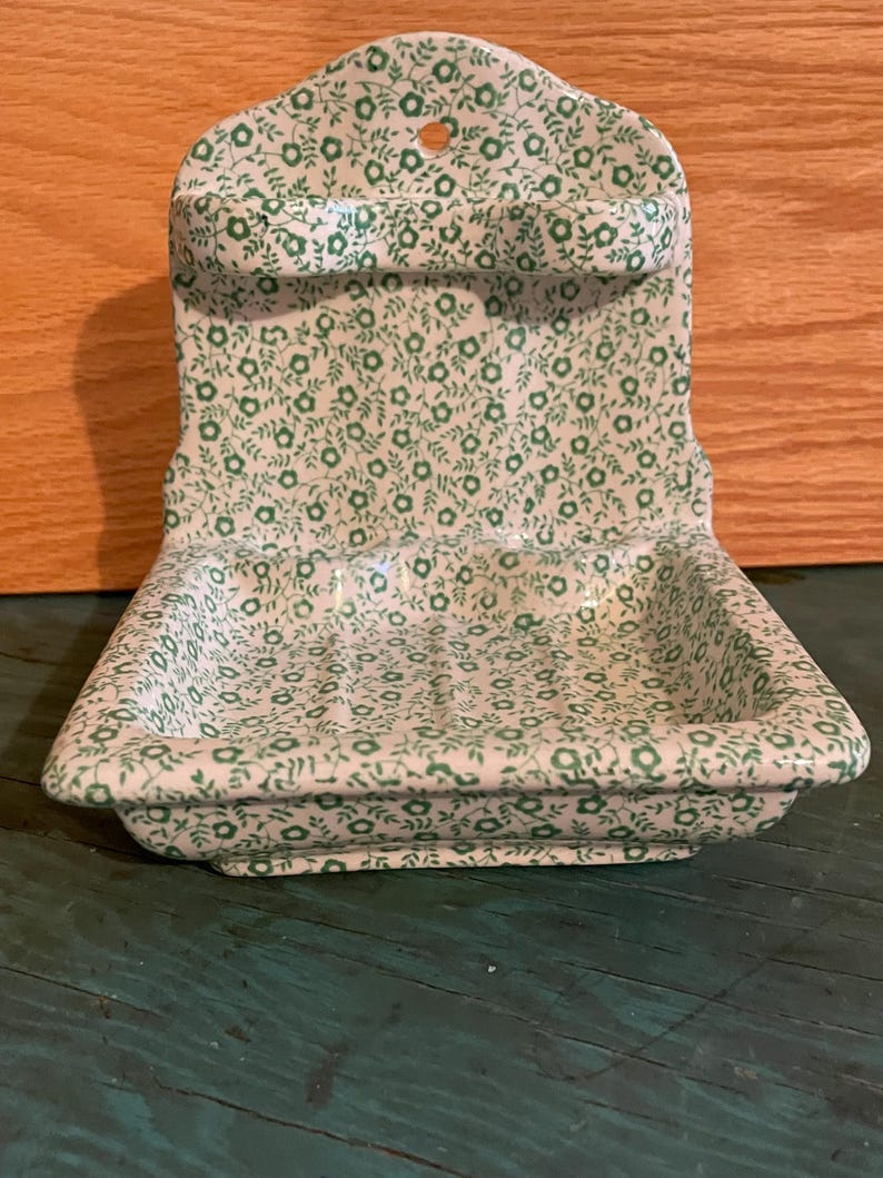 May include: A white ceramic soap dish with a green floral pattern. The dish has a raised edge and a small ledge on the back for hanging. The dish is designed for holding soap bars.