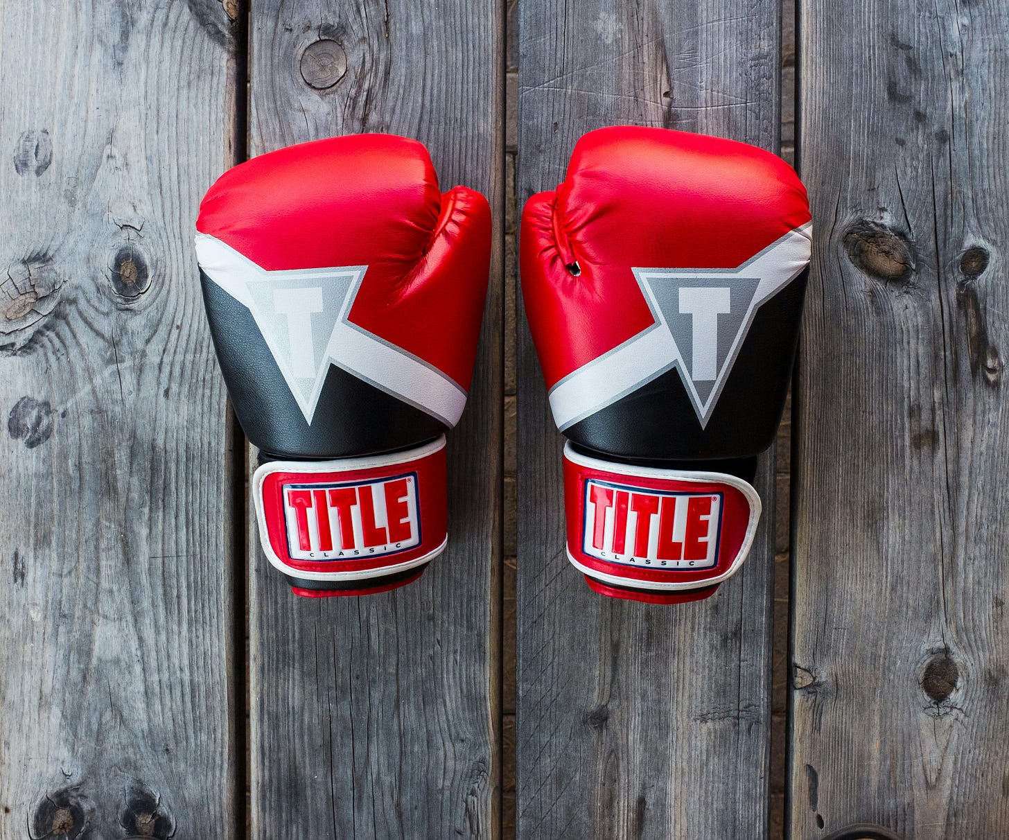 A pair of Red, Black and White Boxing Gloves sit on an ash coloured, wooden tables. You can see the grains and the knots on the wood. The Gloves have the word Title written on the wrist bands