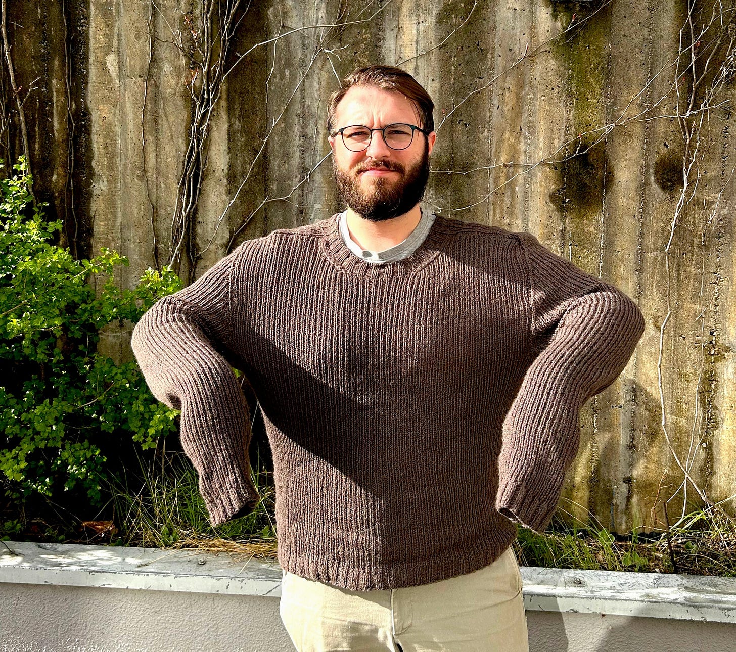 A person wearing a brown 1x1 ribbed sweater, standing against a concrete wall with arms bent. The sleeves are extended over the wrists, indicating excess length.
