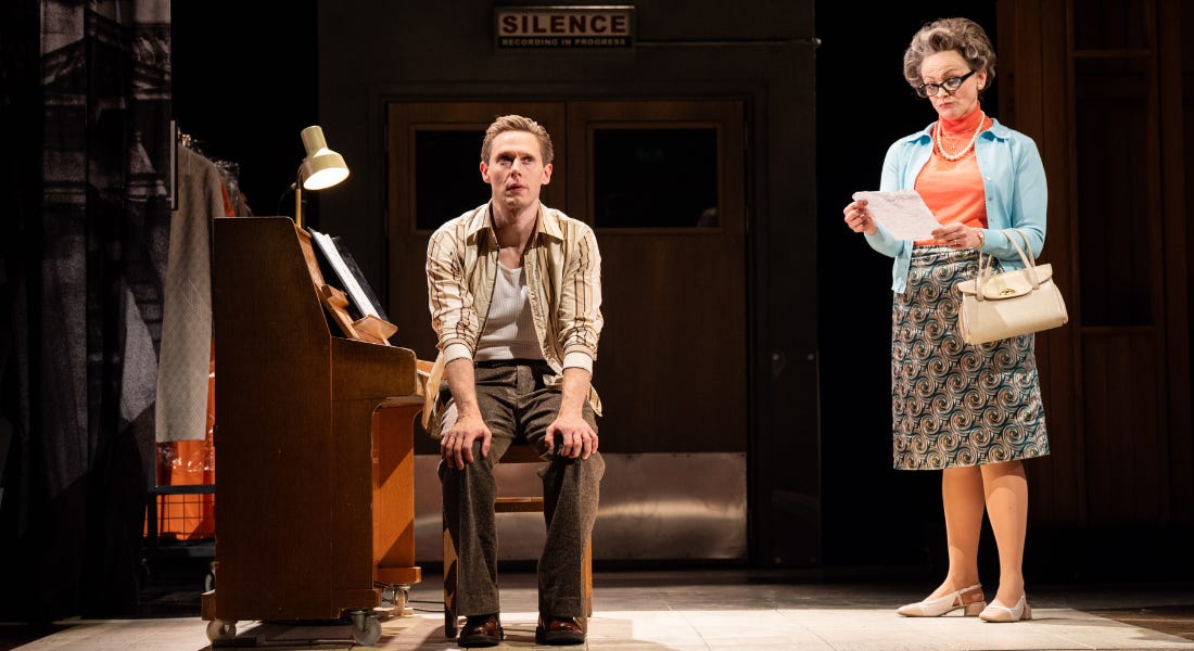 A man in a striped shirt, white vest and brown trousers sits at a piano stool, facing away from the piano and towards the camera. He has his hands on his knees and appears pensive. Behind him to the right is a woman in a blue cardigan, pink rollneck and patterned skirt. She is reading a sheet of paper and has her lips pursed in a disapproving expression.