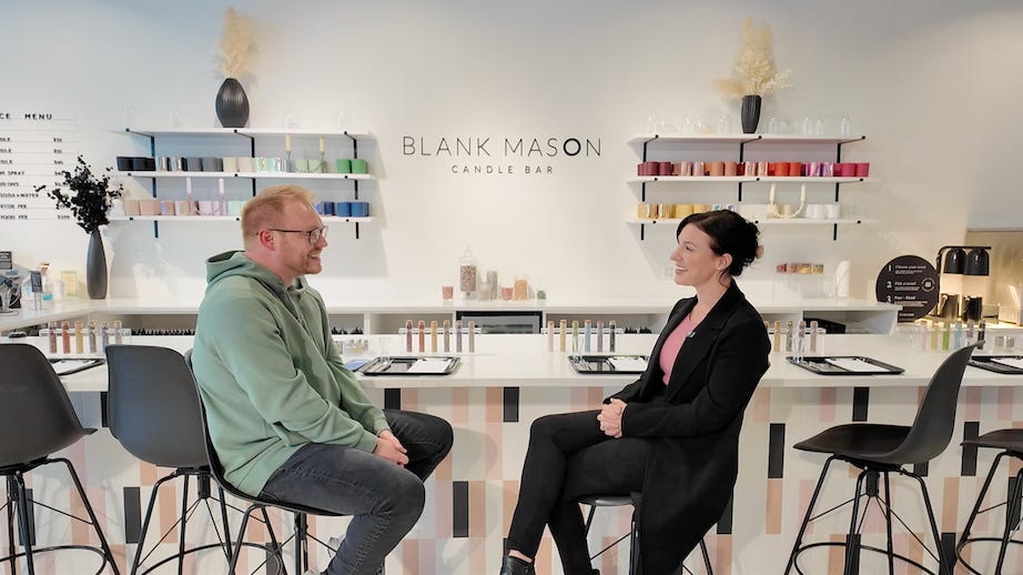 Two people sitting on stools in the Blank Mason Candle Bar. A bright, modern interior with 'BLANK MASON CANDLE BAR' signage visible on the wall behind them. Colorful candle samples are displayed on shelves along the walls.