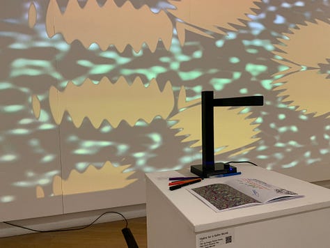 A variety of new media art works using screens, projections, including a large yellow screen and an anaologe cassete with the caption "an actual AI artist" 