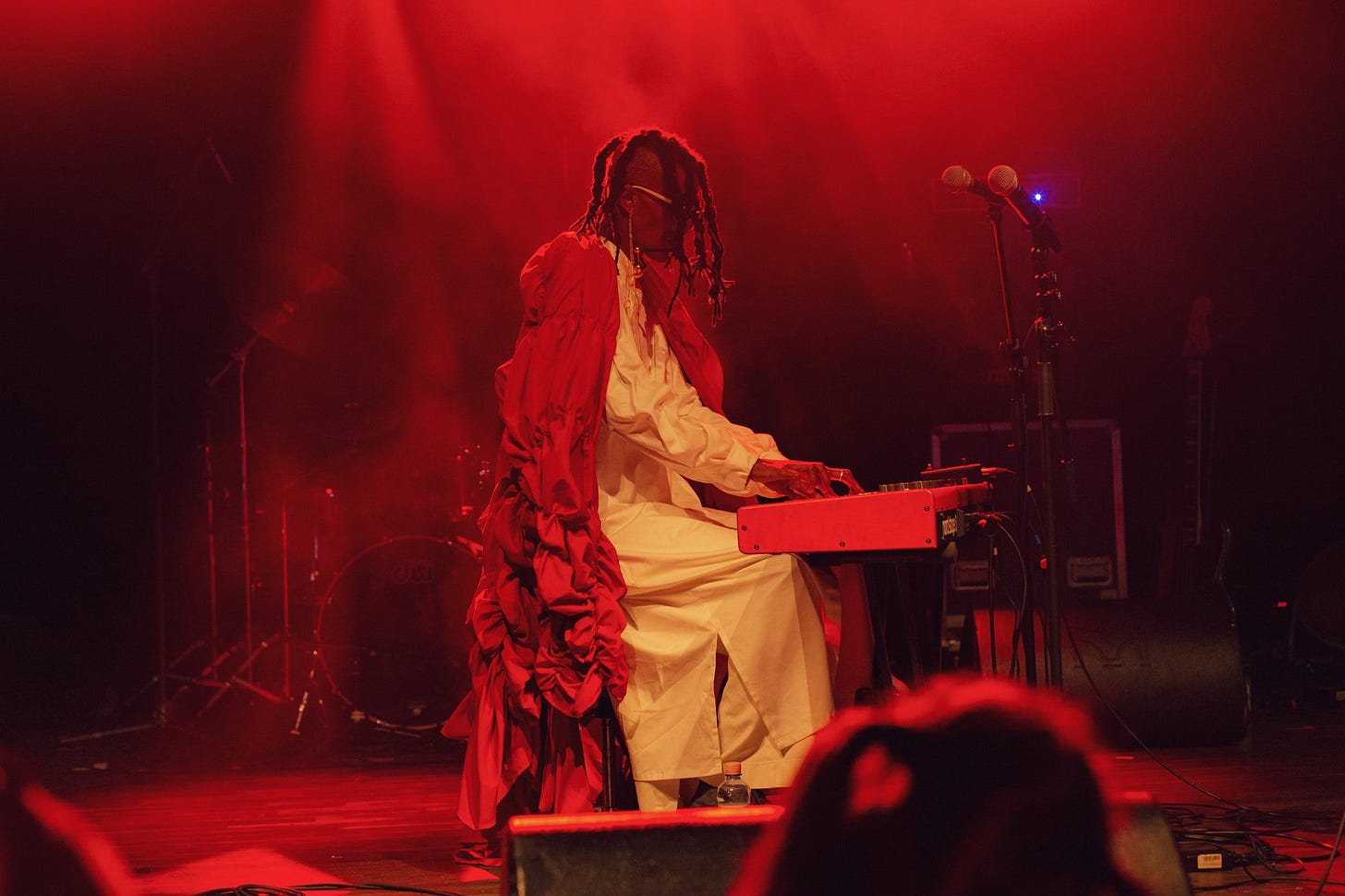 Imagem: Fundo vermelho, artista negro sentado ao piano usando um vestido branco longo e uma capa vermelha.