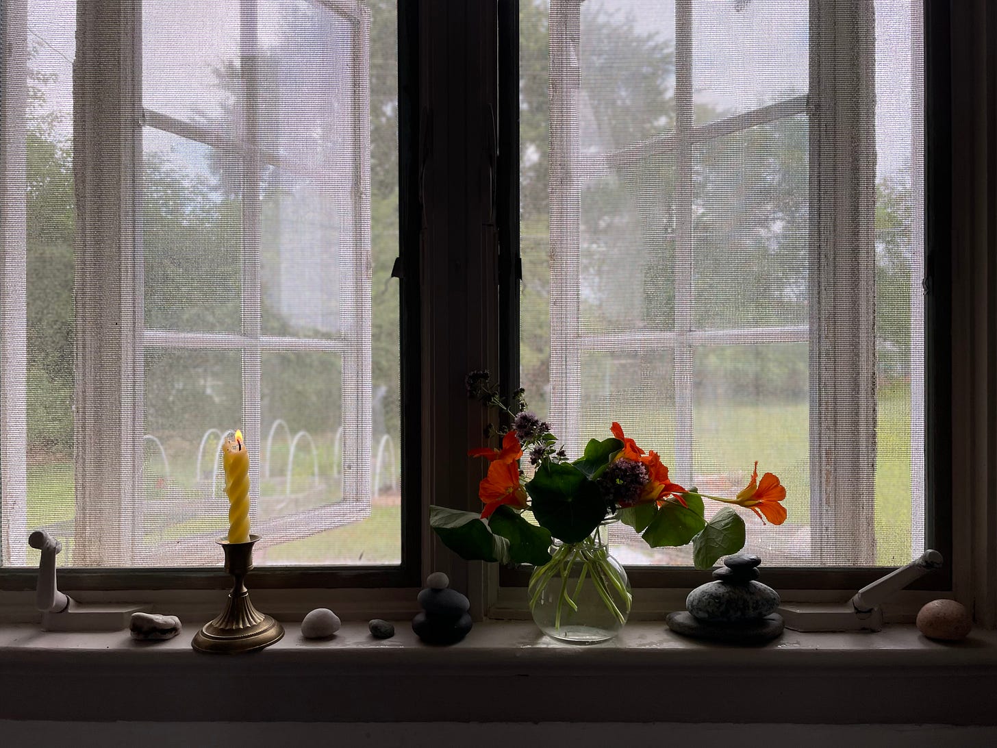 A windowsill in the front the kitchen sink. The windows are the hand cranking sort and they’re open. On the windowsill, which is taupe colored, there are eleven rocks collected from the seaside. Several rocks are stacked on top of each other as cairns. There’s also a beeswax candle that is lit and a glass vase of nasturtiums and oregano blooms.