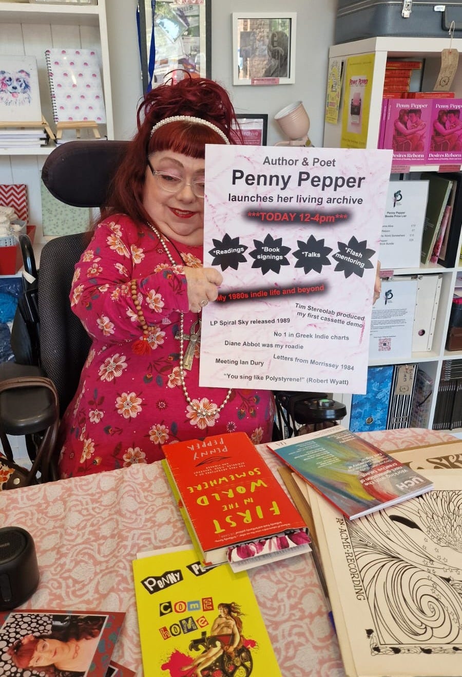 A picture of Penny, who is a wheelchair user and wears glasses, sitting in front of her Living Archive in the shop Crafting Connections in St Leonards-on-Sea. Penny is wearing a bright pink dress with floral detail and has her red hair in a high bun with hairy accessories. She’s surrounded by items from her archive, and her books are on a shelf behind her and on the table in front – copies of First in the World Somewhere, Come Home Alive and her LP Spiral Sky. Penny is holding a poster from an event, which highlights some of her archive material. The light is bright and uplifting and Penny is smiling.