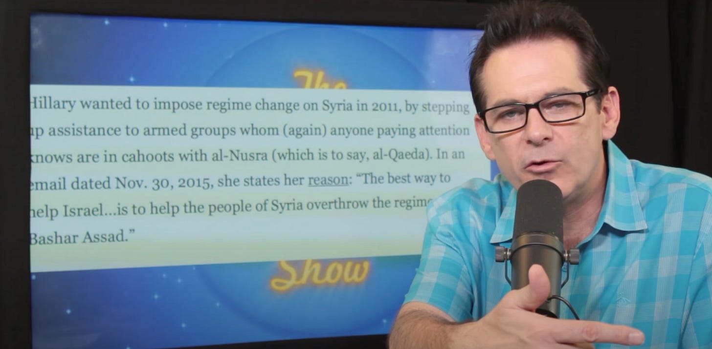 A man with glasses, a blue and white checkered shirt, and a microphone in front of a wall of text.
