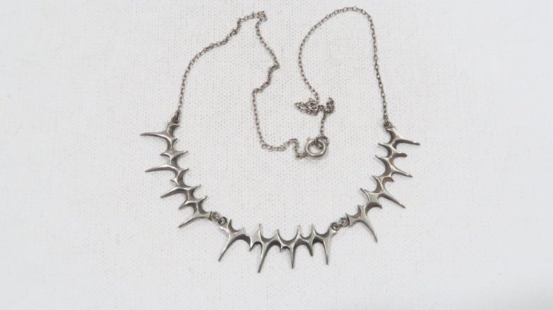 May include: A silver-toned necklace featuring a series of stylized, spiky starburst pendants. The necklace has a delicate chain and a lobster clasp closure. The design is symmetrical and the necklace is displayed on a white surface.
