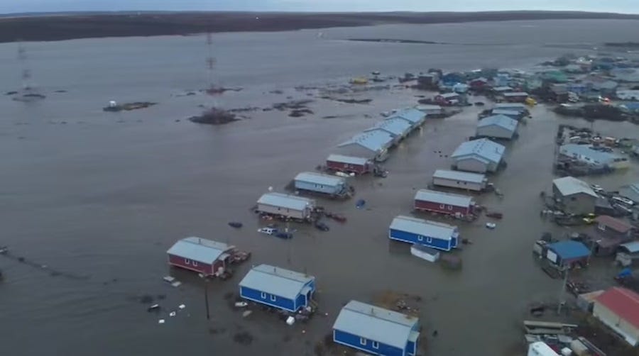 Aerial photo of rows of homes and commercial buildings in a small Alaska village, all surrounded by floodwaters.  Prominent in the foreground are nine prefabricated houses in various colors, and another five, larger prefab warehouses. A mix of other more varied homes and businesses can be seen in the background, they too are surrounded by water. 