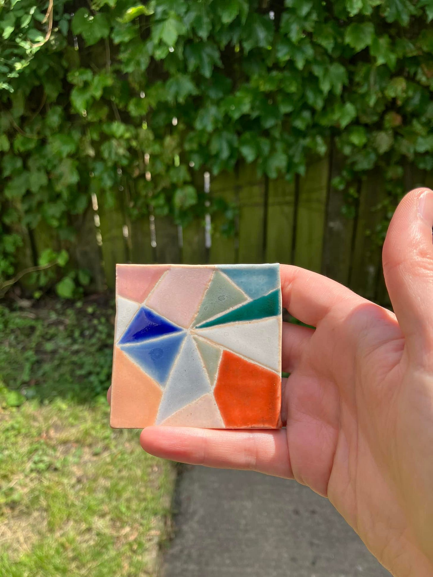 multicolored test tile with geometric design, held up in natural light, in front of an ivy covered fence