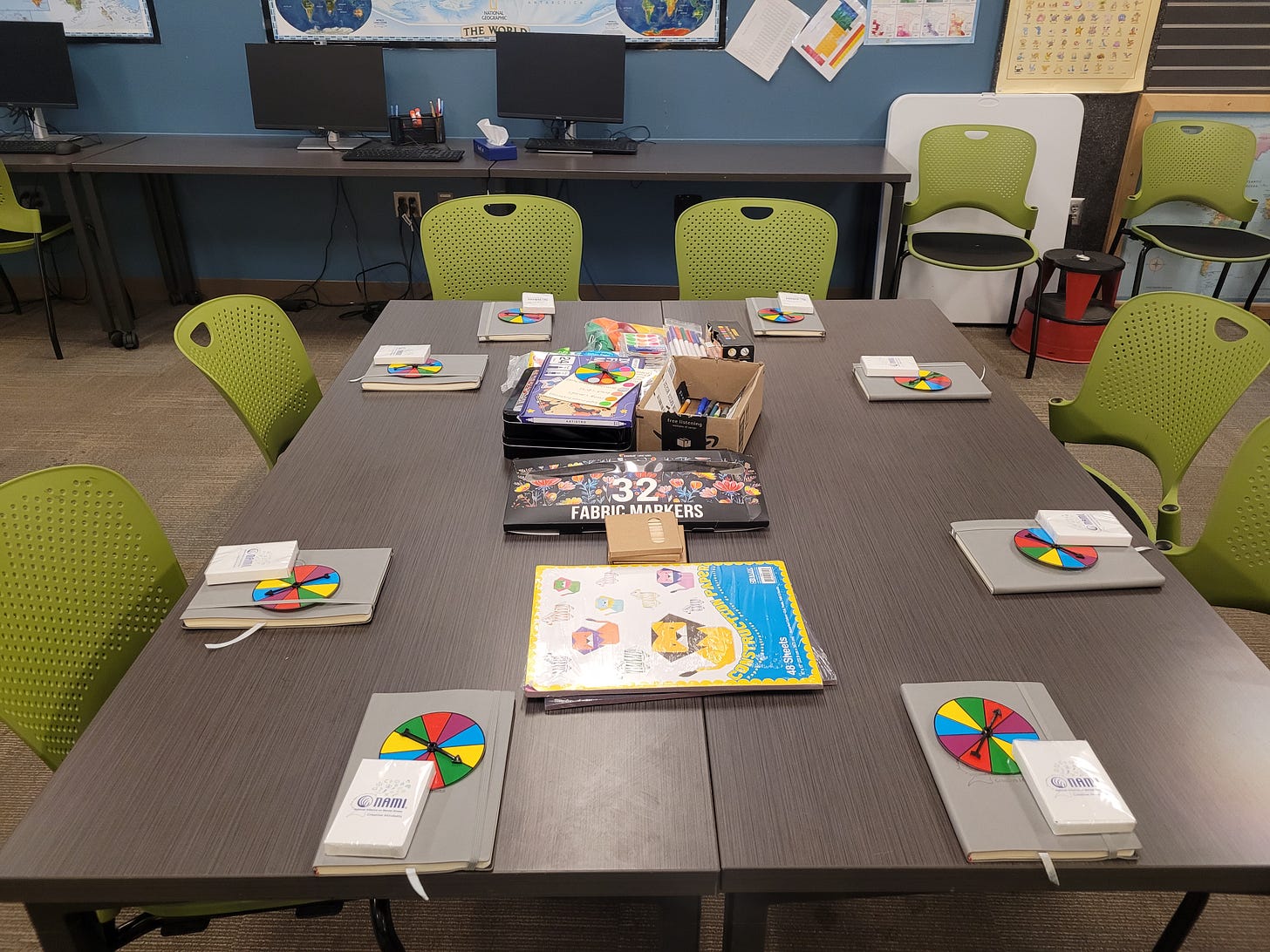A large square table with eight chairs gathered around it. A color wheel, a notebook, and an art supply kit sit in a stack at each spot. Piles of art and craft paper are in the center of the table.