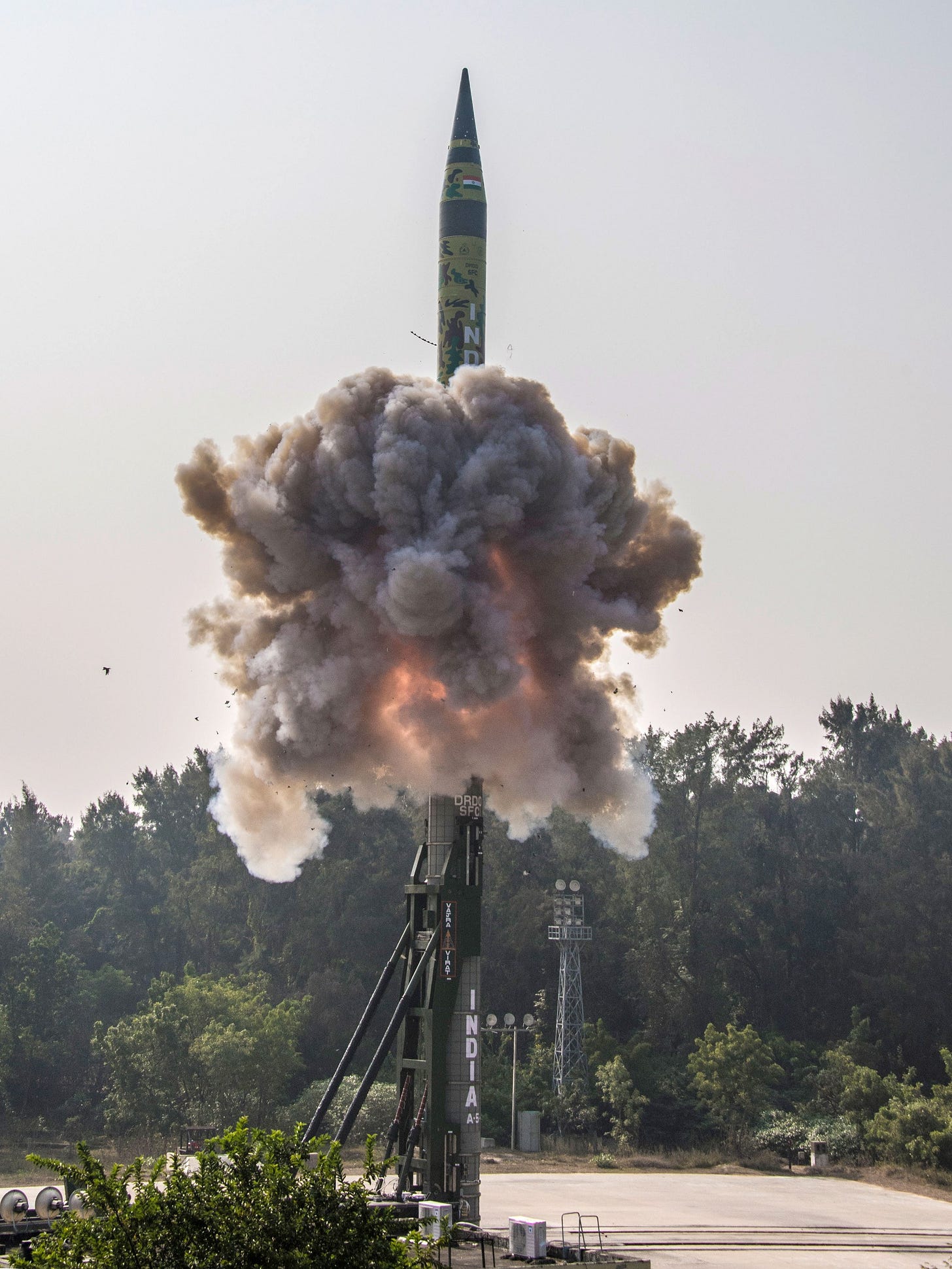 A ballistic missile marked INDIA rises from a canister launcher in a cloud of exhaust, above a treeline