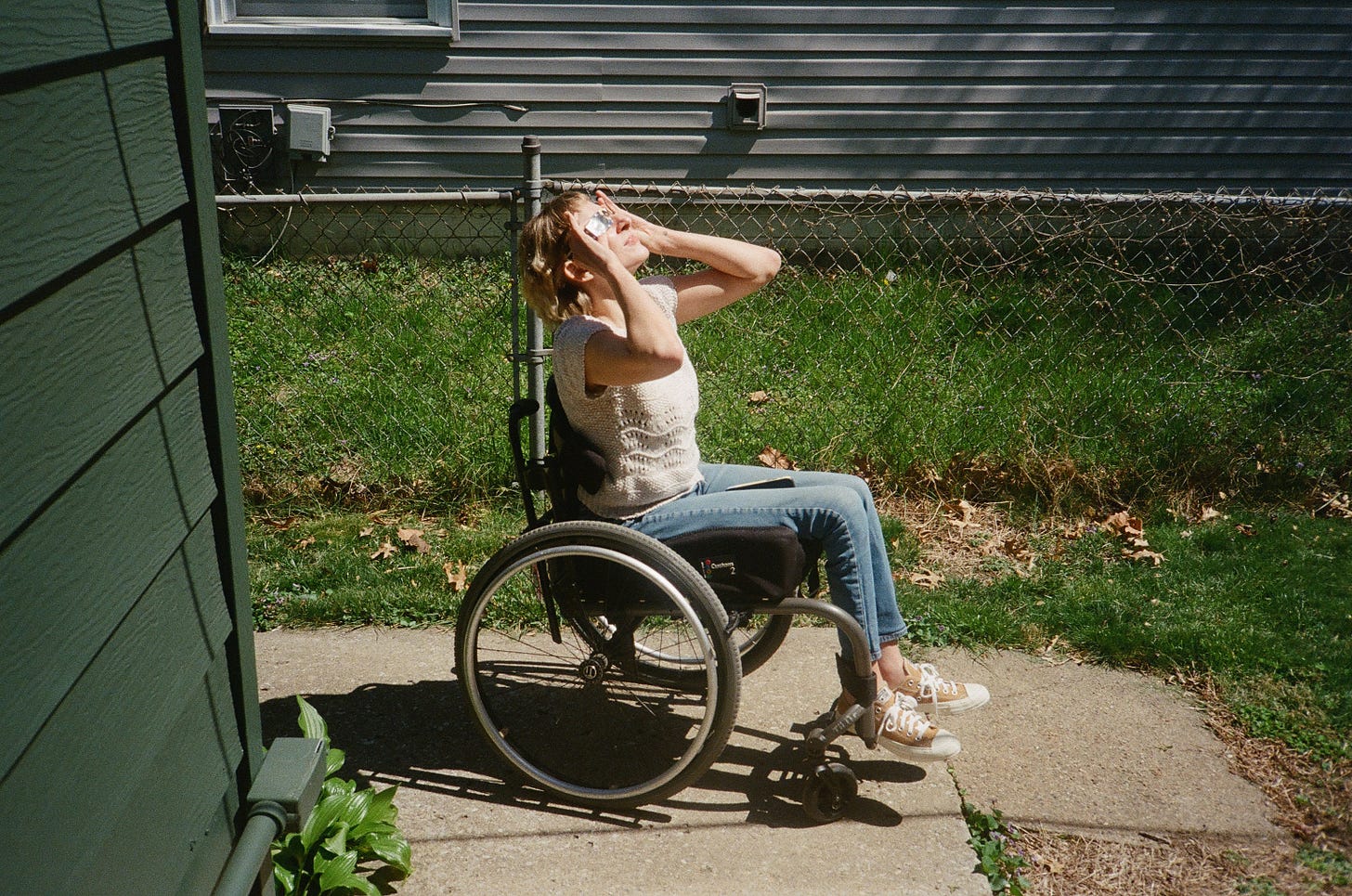 Rebekah sits in her wheelchair with a pair of solar eclipse glasses held up to her eyes, her face tilted toward the sky.