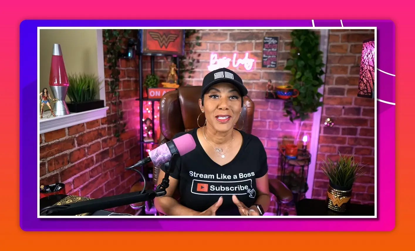 Host smiling with open hands in a colorful home studio, wearing a 'Stream Like a Boss' shirt and a visible pink microphone