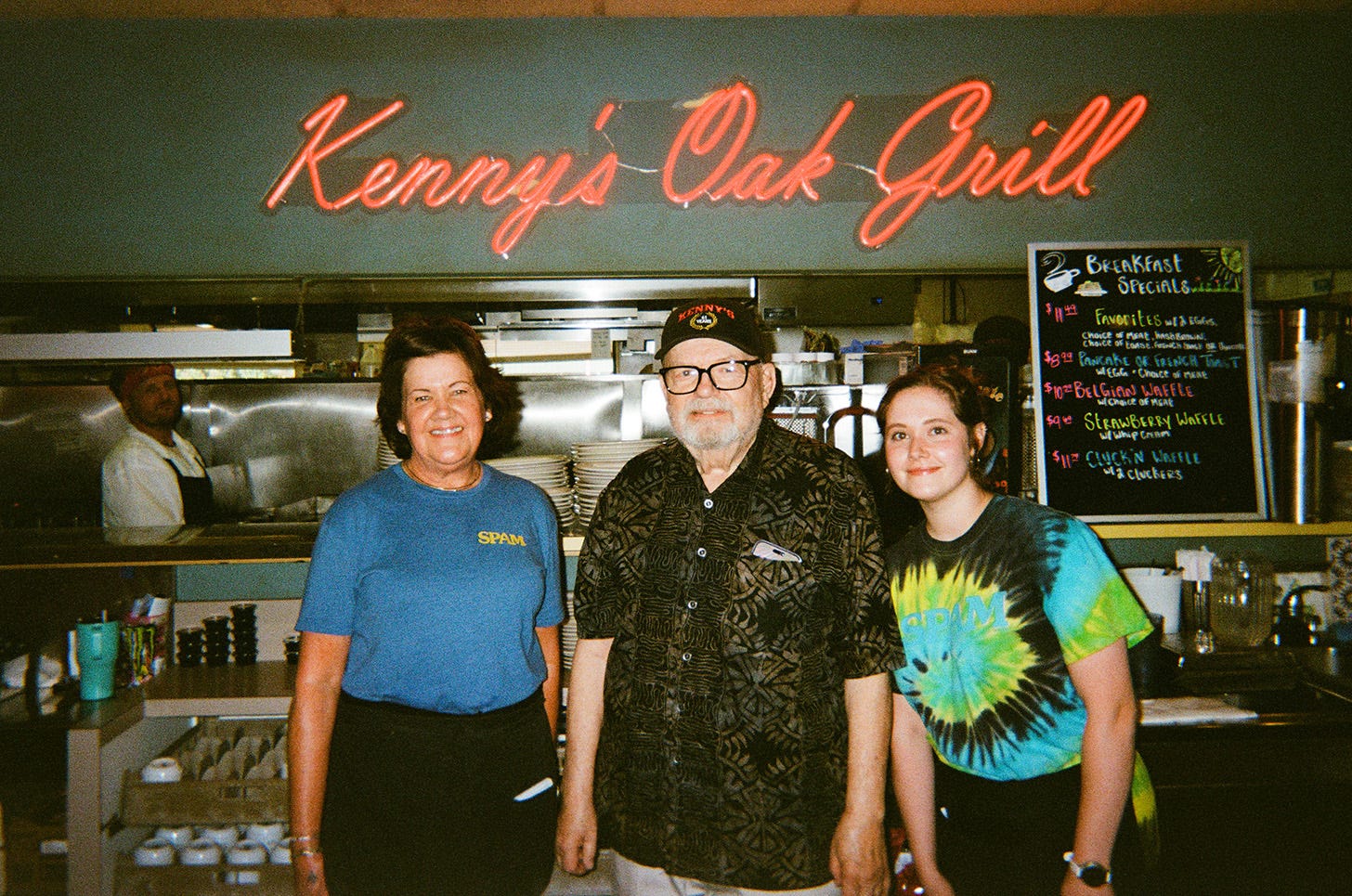 The owner, Kenny, smiles for a photo with two servers. A cursive neon red sign reads, "Kenny's Oak Grill" behind them