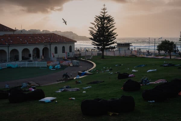 Picnic blankets, lawn chairs and other personal items are strewed across a lawn in front of a low-rise building. A beach is seen in the background.