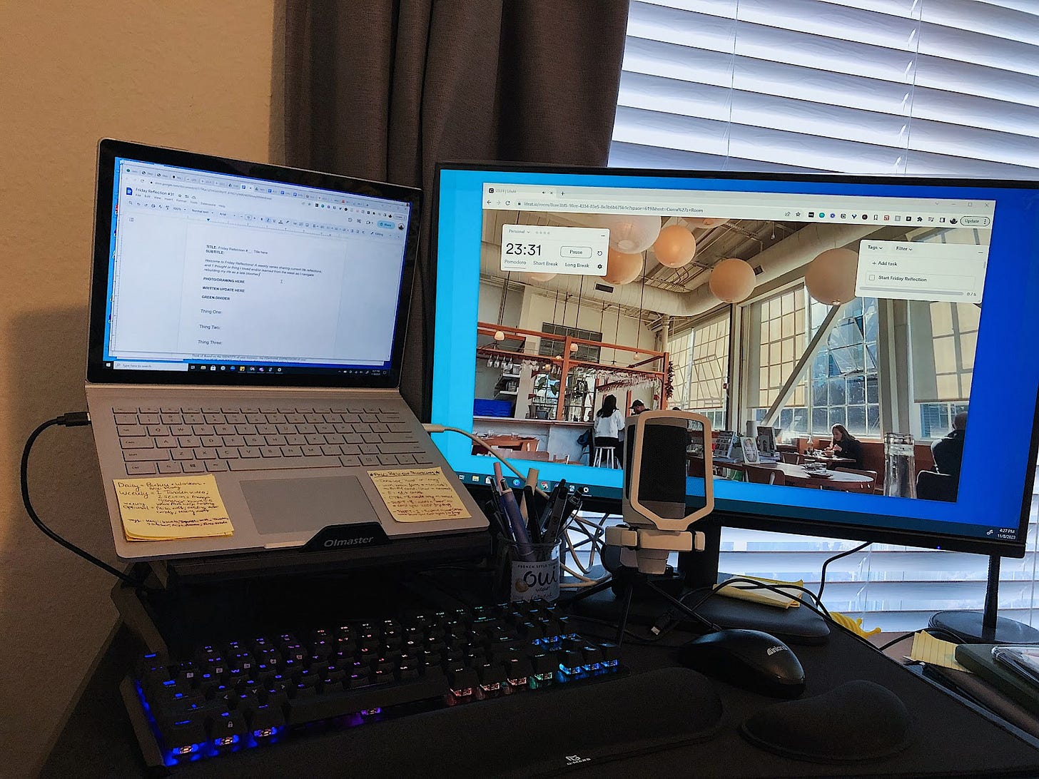 A laptop elevated on a stand with a LED keyboard underneath it, a big second monitor on the right, and a mic in the middle and a black mouse in front of it. On the screen of the second monitor is an ambient and bustling cafe