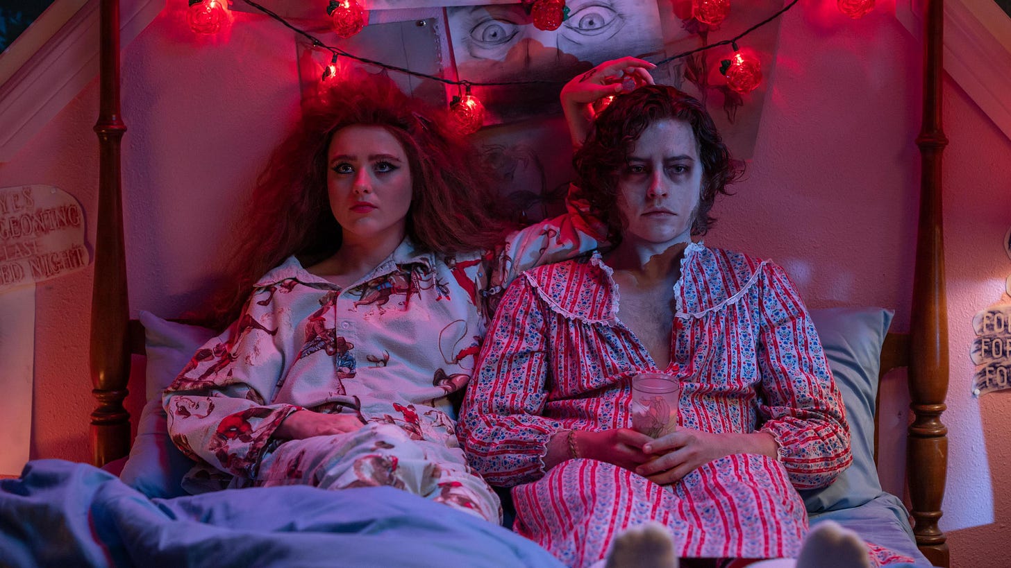 Kathryn Netwon, a young woman with frizzy hair in cowboy-print pajamas, sits in bed next to Cole Sprouse, a zombie in a vintage red and blue sleepgown, under red rose-shaped string lights.