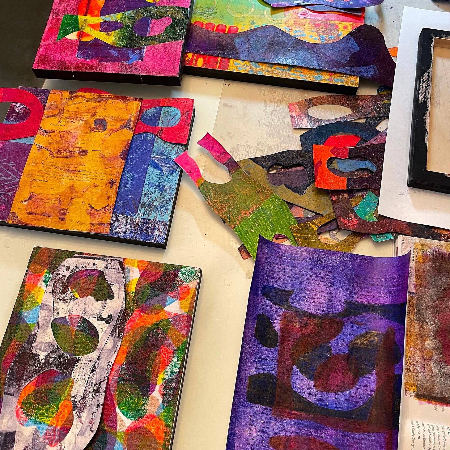 A studio table covered with colorful collage works-in-progress. Brightly painted and printed papers in purples, yellows, greens, and reds are layered and cut into organic shapes. Some pieces are mounted on panels, while others are scattered as scraps, waiting to be arranged.