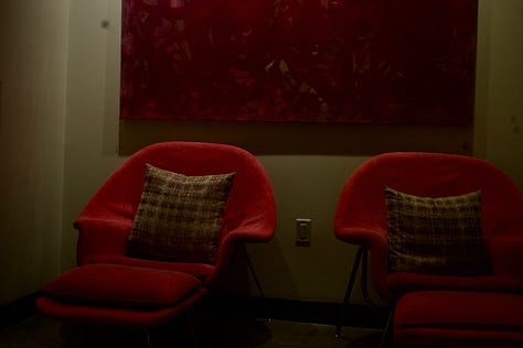 A cinematic series anchored in deep reds and soft shadow: two sculptural red lounge chairs beneath a large abstract canvas; a single orange rose placed against textured wood flooring; a blurred bouquet of roses glowing under streaks of warm light; a nighttime self-portrait reflected in glass doors; and a tightly framed cluster of red roses resting on a neutral surface. The compositions emphasize symmetry, negative space, reflection, and saturated color, creating a quiet narrative of intimacy, interior space, and becoming.