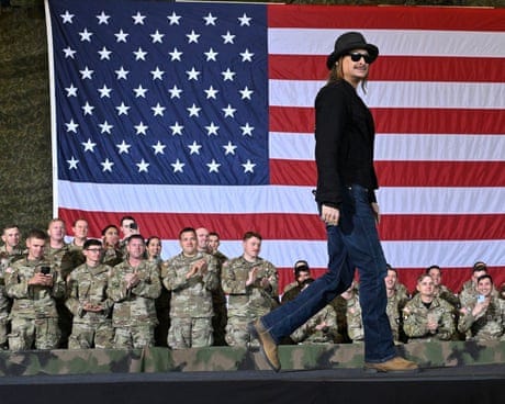 a man in a hat walks on stage backed by a giant us flag and troops in uniform