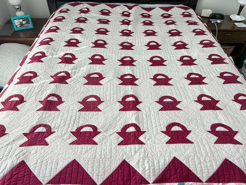 May include: A white quilt with a burgundy basket pattern. The quilt is made with a diamond pattern and has a quilted texture. May include: A white quilt with a burgundy basket pattern. The quilt is made with a diamond pattern and has a quilted texture.
