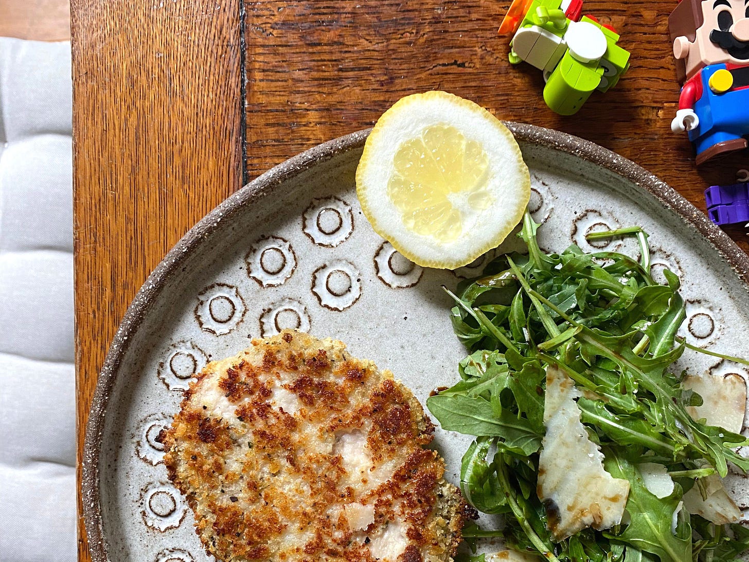 A plate on a wooden table top. Plate holds a breaded piece of meat, rocket salad and lemon slice. Lego Mario is also on the table.