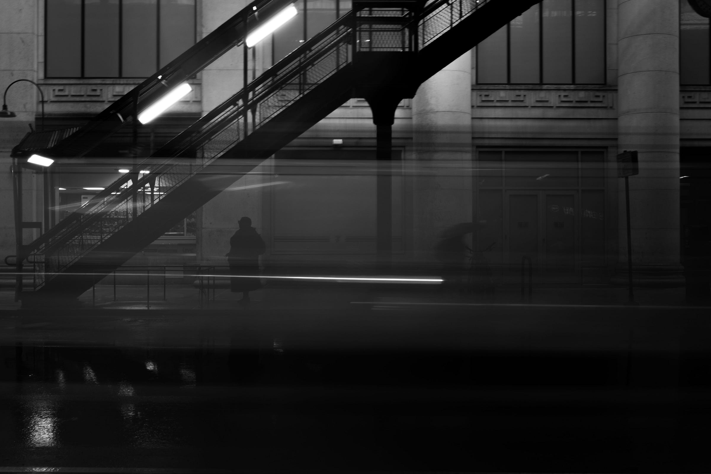 A shadowy figure stands beneath an elevated train staircase at night, while blurred light streaks from passing traffic create a ghostly, cinematic effect on the wet street. A shadowy figure stands beneath an elevated train staircase at night, while blurred light streaks from passing traffic create a ghostly, cinematic effect on the wet street.