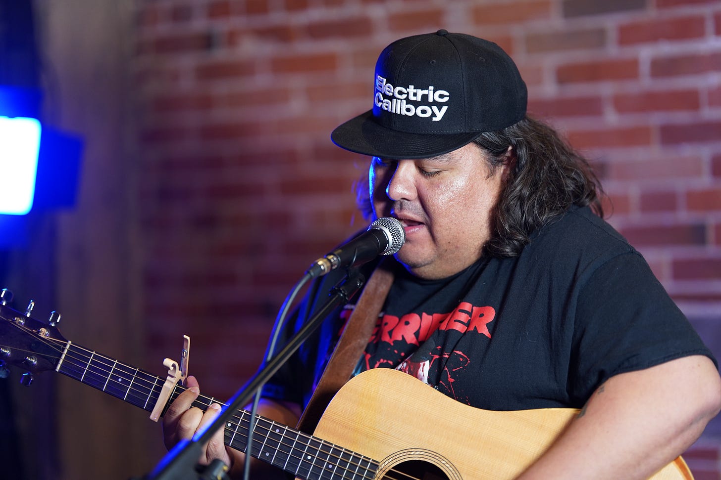 A person standing in front of a microphone, singing. He holds a wooden guitar. 