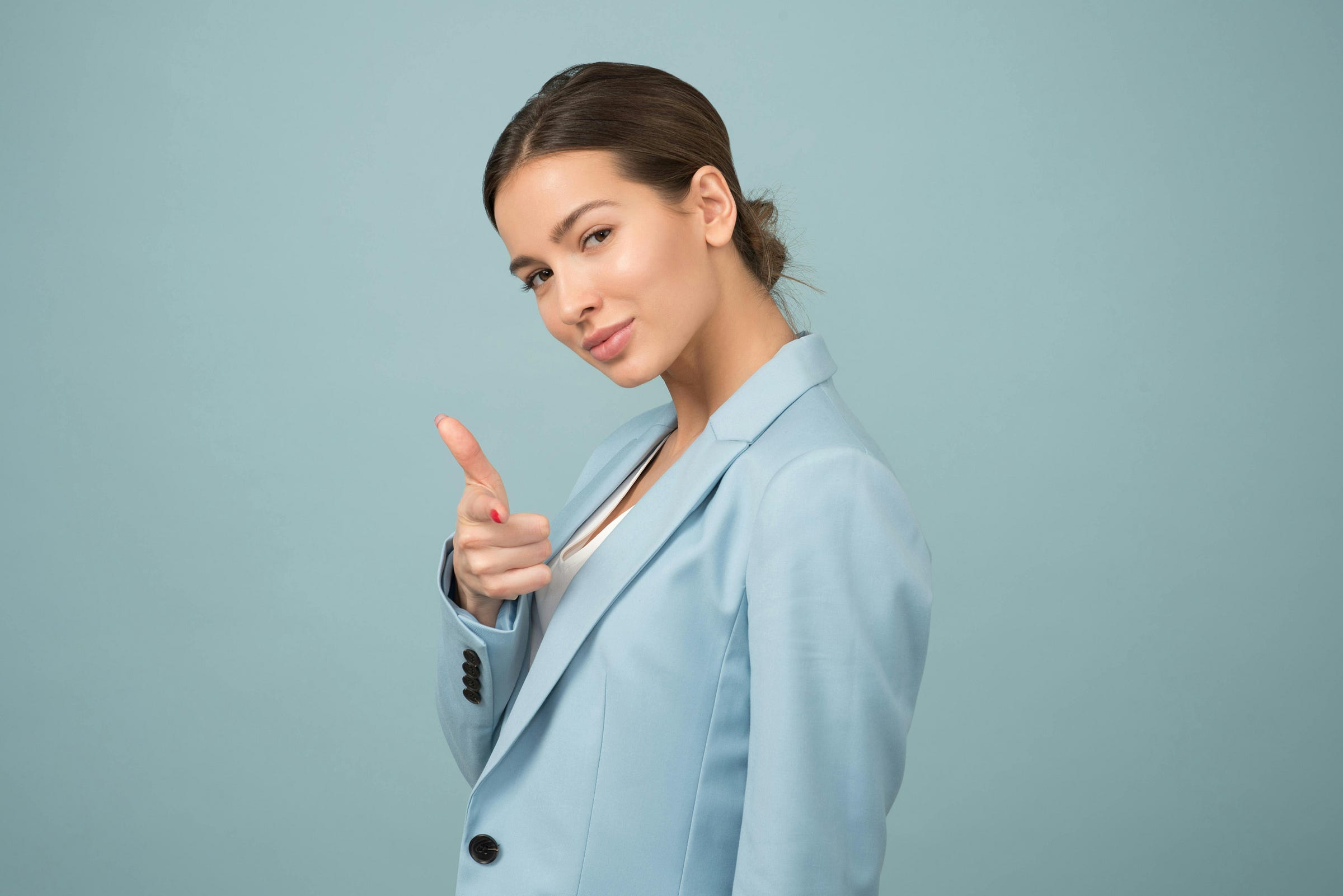 A medium close-up studio shot of a woman with her hair pulled back, wearing a light blue blazer over a white top. She is looking at the camera and pointing her right index finger forward, as if gesturing to the viewer. The background is a solid light blue. A medium close-up studio shot of a woman with her hair pulled back, wearing a light blue blazer over a white top. She is looking at the camera and pointing her right index finger forward, as if gesturing to the viewer. The background is a solid light blue.