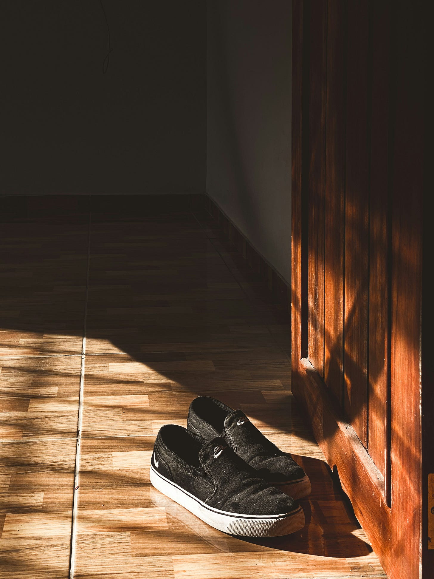 A pair of black slip-on shoes sitting on a wooden floor near an open door, with sunlight casting shadows around.