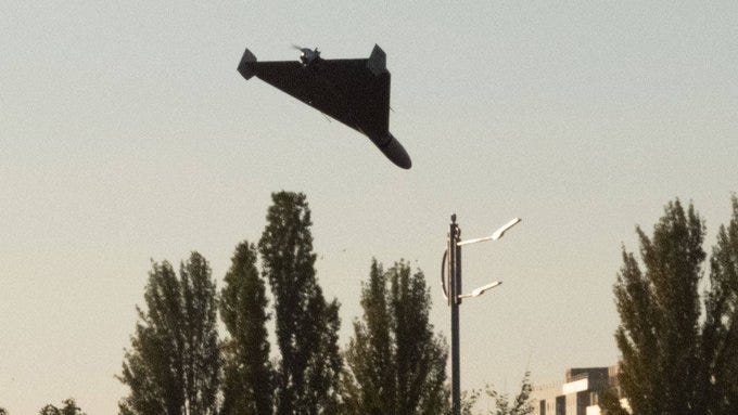 A dark-colored KROOK-1 drone in flight against a clear sky. The drone has a triangular shape with a visible pulsejet engine. Trees and a streetlight are visible below. A dark-colored KROOK-1 drone in flight against a clear sky. The drone has a triangular shape with a visible pulsejet engine. Trees and a streetlight are visible below.