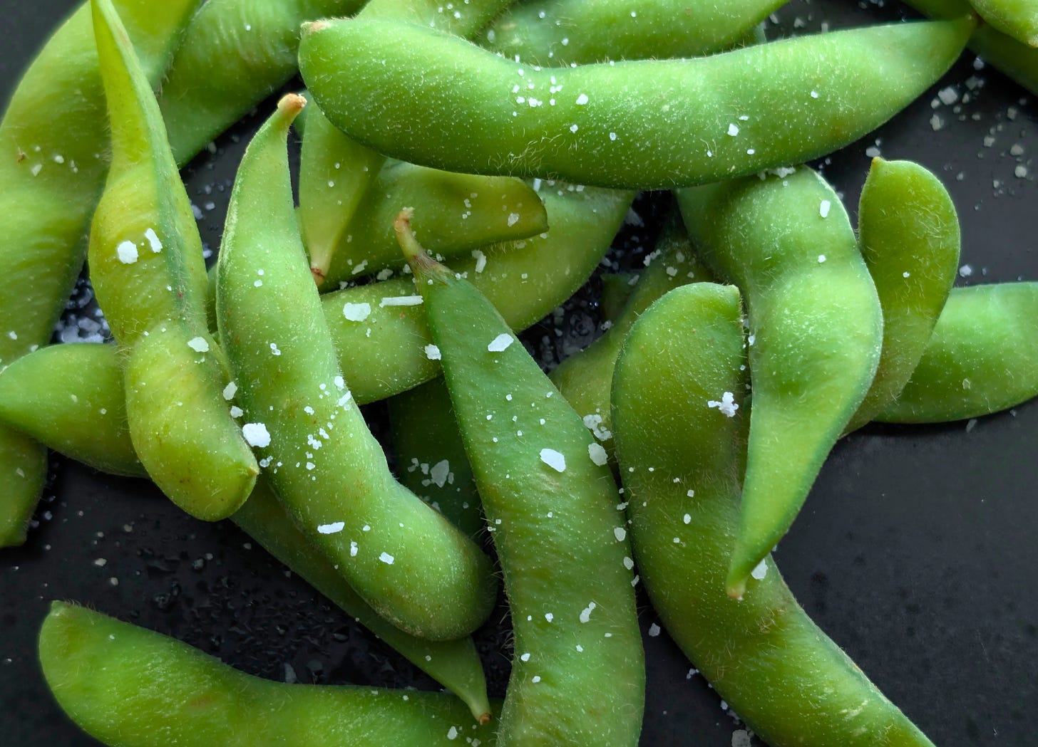 Close-up of cooked edamame pods sprinkled with flaky salt on a black plate.