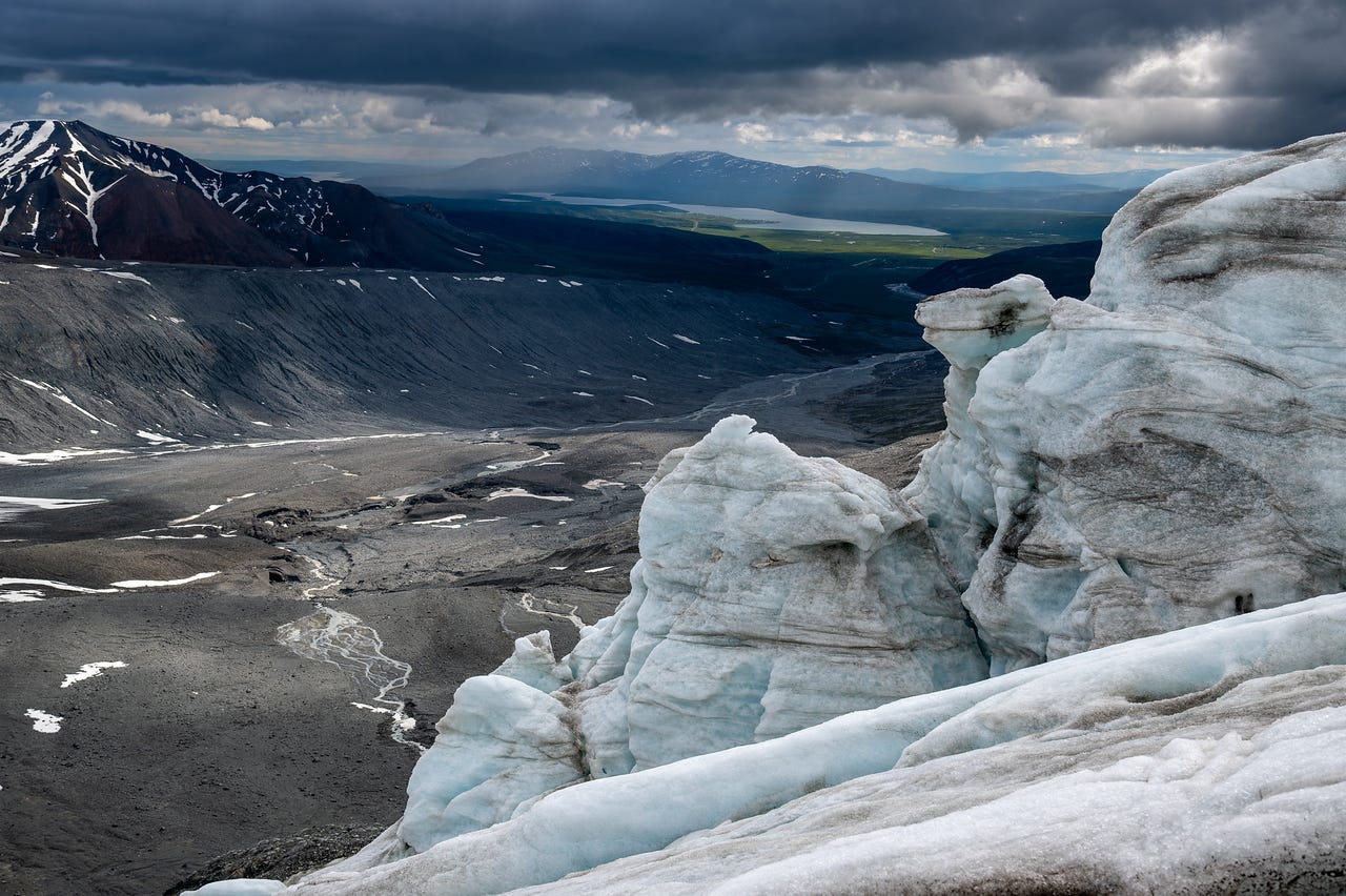From high on the icefall, sculpted towers of blue-white ice stand in the right foreground while the view drops to a vast grey outwash valley, where a braided stream winds over gravel between dark lateral moraine walls. Far beyond, sunlight breaks through storm cloud onto green lowlands and the pale sheet of Summit Lake, with rounded mountains on the horizon under a shelf of nearly black sky.
